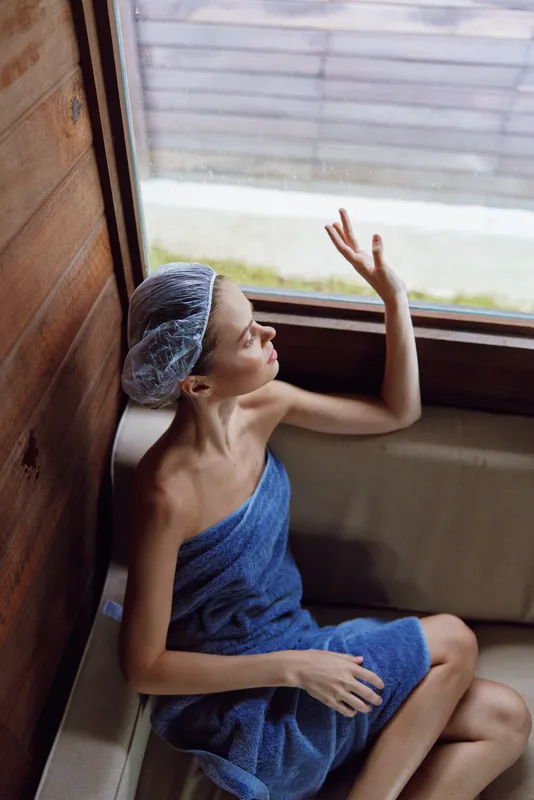 young woman relaxing in sauna wrapped in blue towel and wearing shower cap, sitting on wooden bench and enjoying warm steam indoors