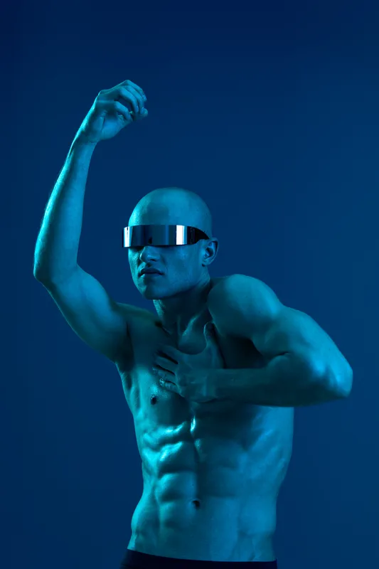 Dynamic man striking a pose with futuristic sunglasses, showcasing strong physique in a captivating blue light. A perfect blend of art and fashion for modern visuals.