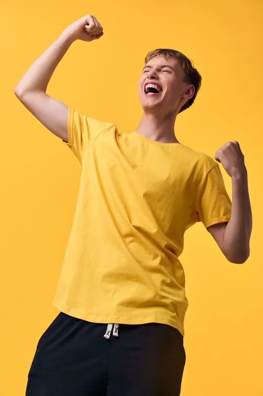 Joyful man in a bright yellow t shirt celebrates a win with raised fists against a vivid orange backdrop, conveying energy, enthusiasm and triumph in a dynamic studio scene.