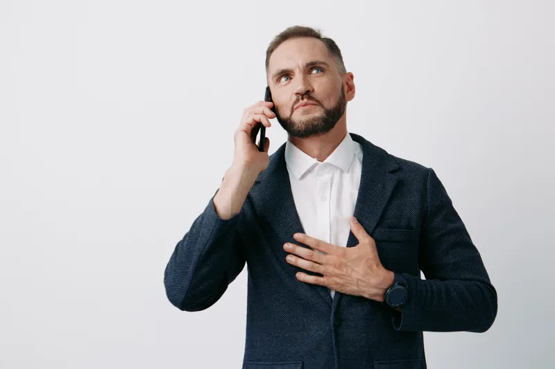 A professional businessman on a call, wearing a dark blazer against an isolated solid color background. He gestures to his chest, signaling confidence and focus while discussing important matters.
