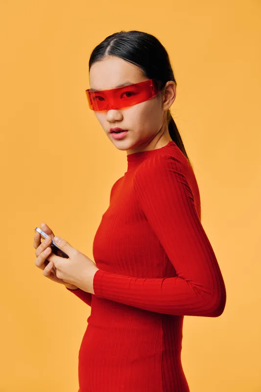 Young woman wearing red turtleneck sweater and futuristic red eyewear looks sideways holding smartphone with both hands on yellow background in studio lighting fashion concept.