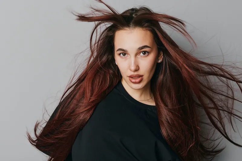 Woman with long flowing hair and intense gaze wearing black shirt on gray background. Portrait of confident female model with natural makeup and dynamic hair movement in studio.