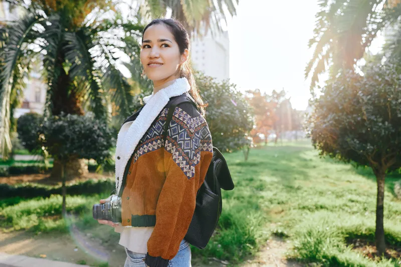 Woman with camera and backpack in a park, wearing a jacket and scarf, enjoying outdoor sunlight and nature during daytime, ready for street photography and exploration