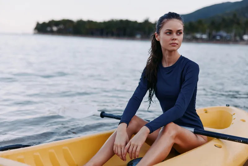 A woman sits in a bright yellow kayak on calm waters, wearing a navy long-sleeve top, with a thoughtful gaze toward the distance and horizon.