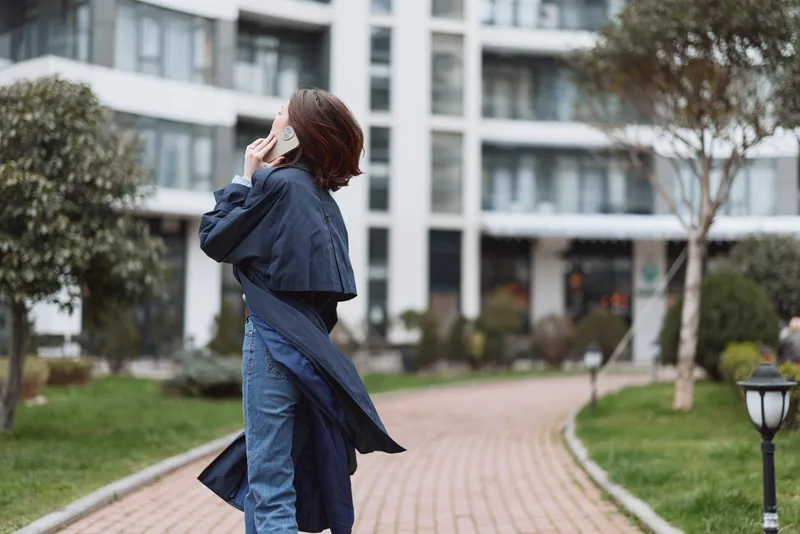 Young woman with short brown hair wearing a long dark coat and jeans walks along a paved path while talking on a smartphone in an urban residential area.