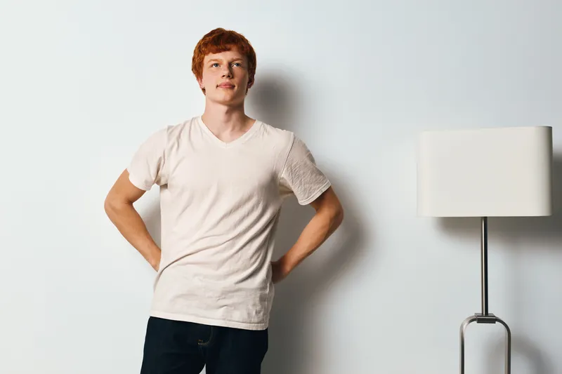 Young man with red hair and fair skin standing confidently in white t shirt and jeans near floor lamp against white wall in bright indoor space with casual pose and relaxed atmosphere