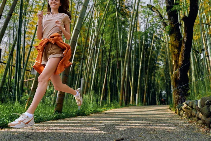 Woman jumping on forest path in bamboo grove, smiling in casual outfit with tied jacket and sneakers, carefree motion and active outdoor leisure in green nature scenery.