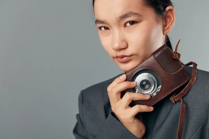 Young woman holding vintage camera close to face with leather case wearing dark blazer against plain background for photography, retro style, and portrait concept in studio setting.