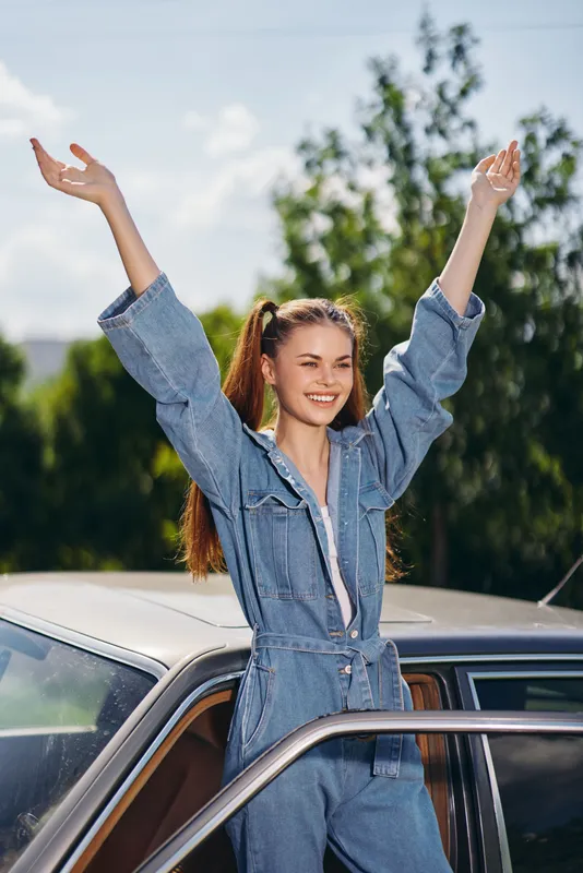 Young woman celebrating joyfully in denim outfit with raised arms, standing by a classic car under a blue sky with trees.