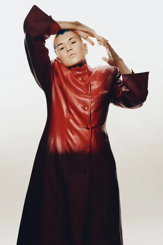 Woman wearing a long red leather coat posing confidently against a light background, showcasing modern fashion and edgy style with a serious expression.