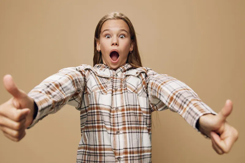 Girl with long hair wearing plaid shirt shows thumbs up gesture with surprised face expression on neutral background. Youth, casual style and positive reaction for success or approval.