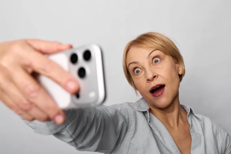 Woman surprised with smartphone selfie, communication and video taking, hand extended, portrait on gray studio background, close up face and emotion.