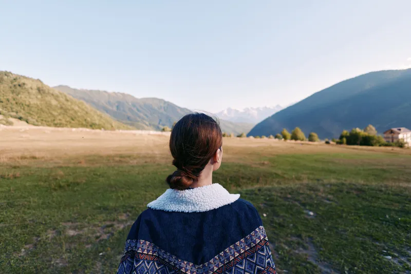 Woman back overlooking mountain landscape and meadow in a valley, outdoors travel scene with calm nature, rural field and distant hills for peaceful exploration and adventure.