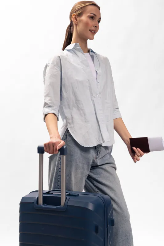 Young woman in casual outfit holding passport and boarding pass stands next to blue suitcase. Travel concept with preparation, journey, and vacation planning on white background.