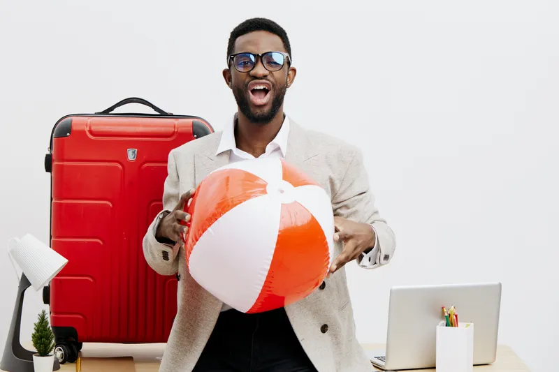 Confident smiling man with beard and glasses holding summer beach ball, standing in front of red suitcase, laptop, and travel accessories on white background. People travel vacation concept