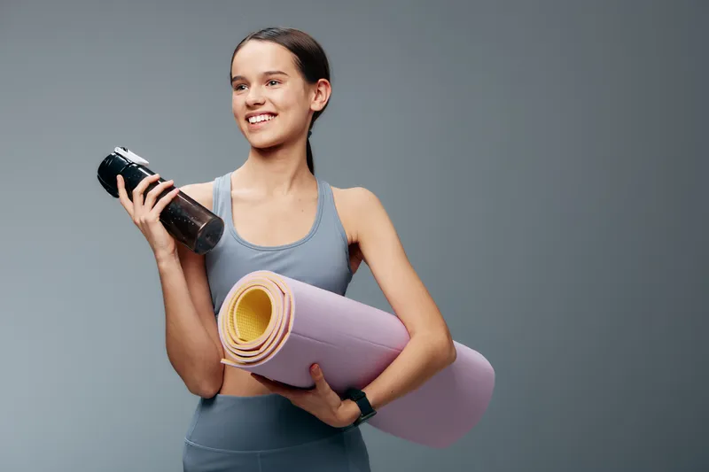 Fitness woman holding a water bottle and rolled yoga mat, ready for a workout. Athletic pose in a simple studio with clean backdrop, showcasing sport, training, yoga, and equipment for a healthy