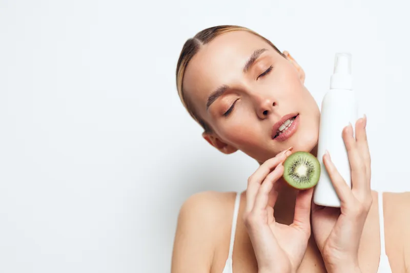 Young woman with clean skin holding a spray bottle and a kiwi fruit, eyes closed, fresh natural skincare concept, white background, minimalistic beauty routine, soft lighting