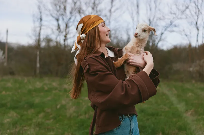 Smiling woman in a brown jacket holds a curious goat kid outdoors in a field, showing a warm lifestyle moment, affection and interaction between human and animal. The soft light and open meadow