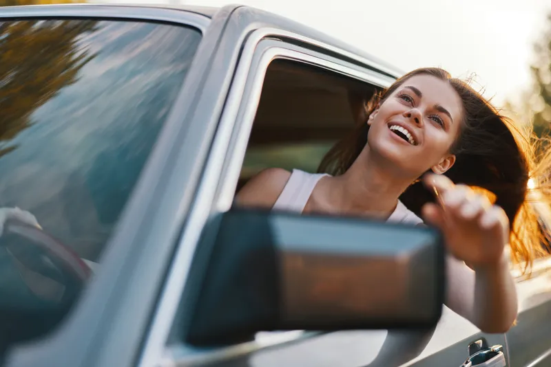 Happy woman enjoying a road trip with her hair flowing in the wind, showcasing a sense of freedom and adventure in a car during a sunny day.