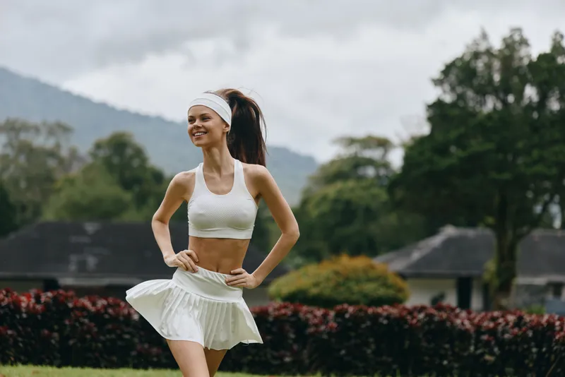 Happy young woman exercising outdoors in a white sports outfit, enjoying fitness and health in a lush green park during a cloudy day