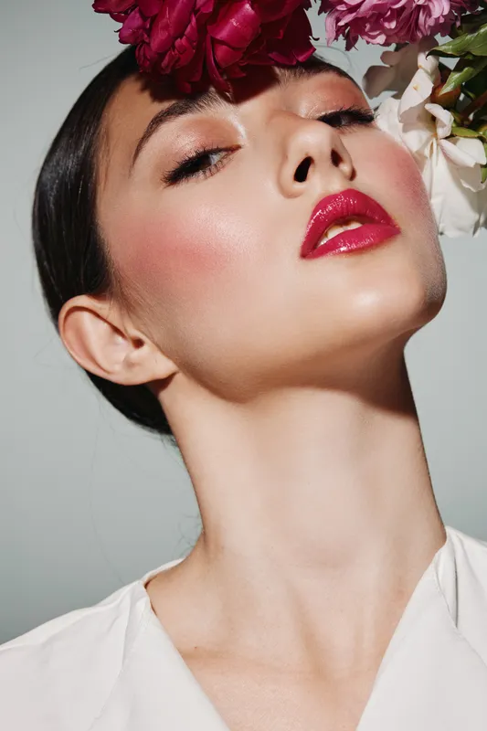 Close-up of young woman with glossy red lips and shimmery eyeshadow posing with flowers on her head. High fashion beauty look with creative floral accessory in studio lighting.