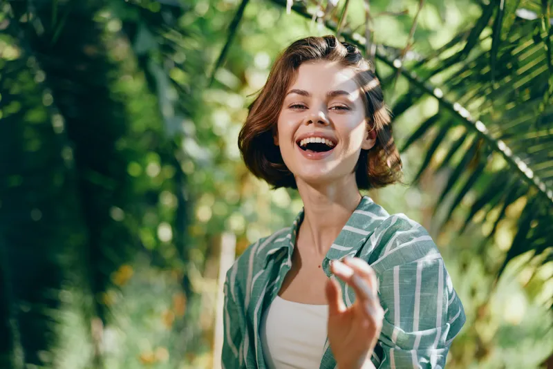 A joyful woman outdoors smiles brightly, enjoying sunny weather. She wears a striped green shirt and stands among lush trees, radiating happiness and carefree energy.