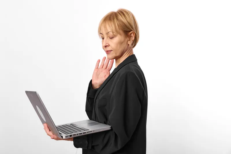 Senior woman with laptop in office setting, technology communication concept, concerned expression, hand near face, typing on screen, white background