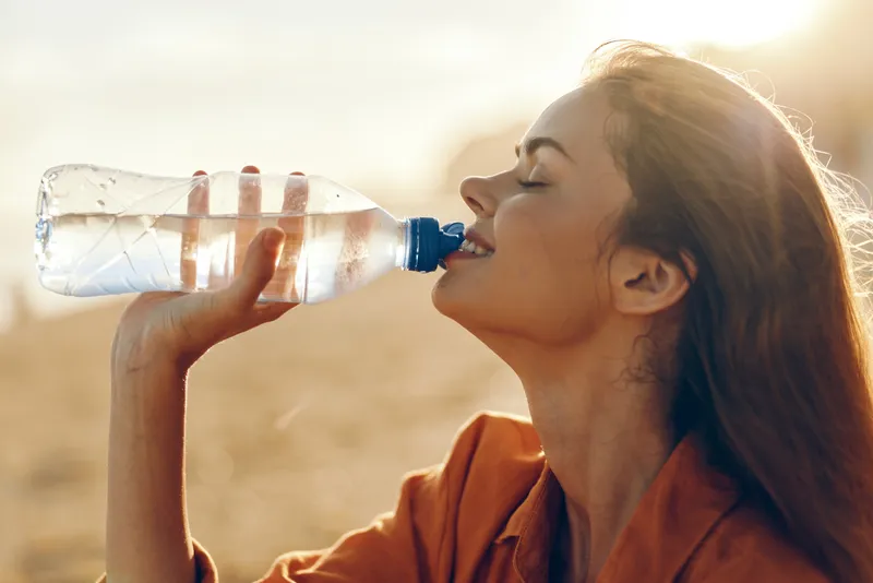A woman in casual wear drinks water from a bottle outdoors on a sunny day, highlighting hydration, stamina, and a healthy lifestyle in an active moment.