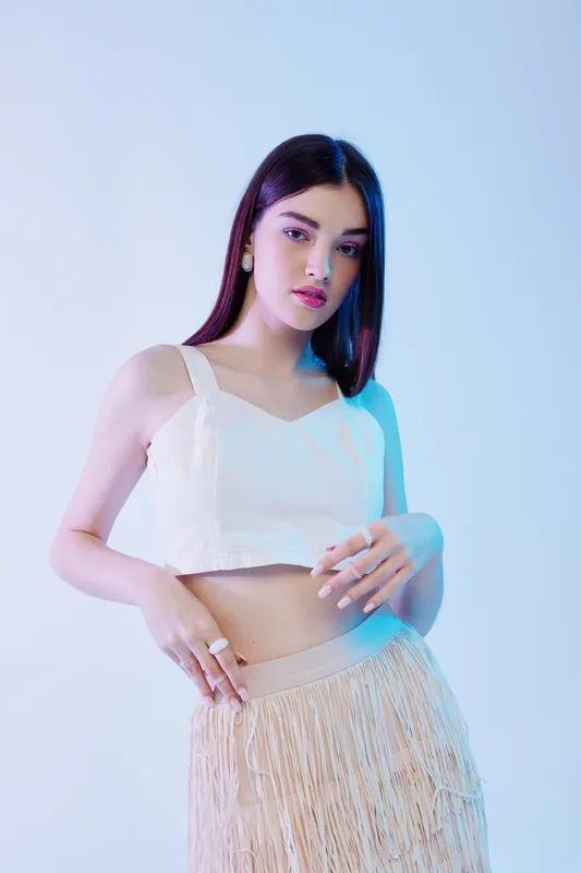 Young woman in a white crop top and beige fringe skirt poses confidently against a light background. Concept of woman beauty fashion, style, and elegance.