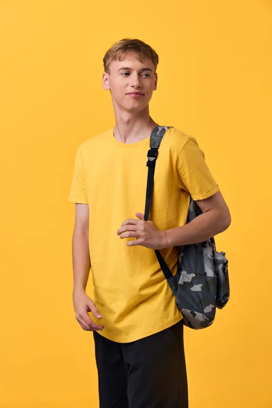 Young student wearing a yellow t shirt with a camouflage backpack stands against a bright backdrop in studio light, casual pose suggesting readiness for class, travel, and daily activities