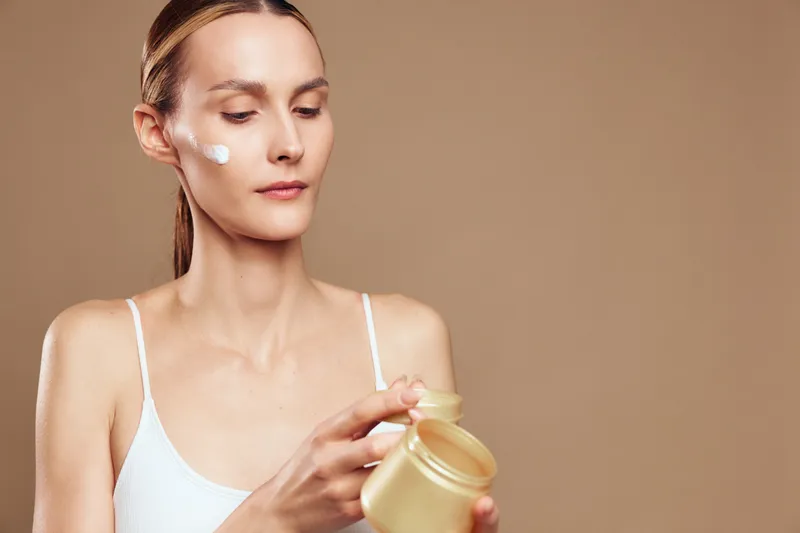 Young woman with clear skin applies face cream from jar on beige background, skincare routine concept, natural beauty, focused expression, wellness and self care