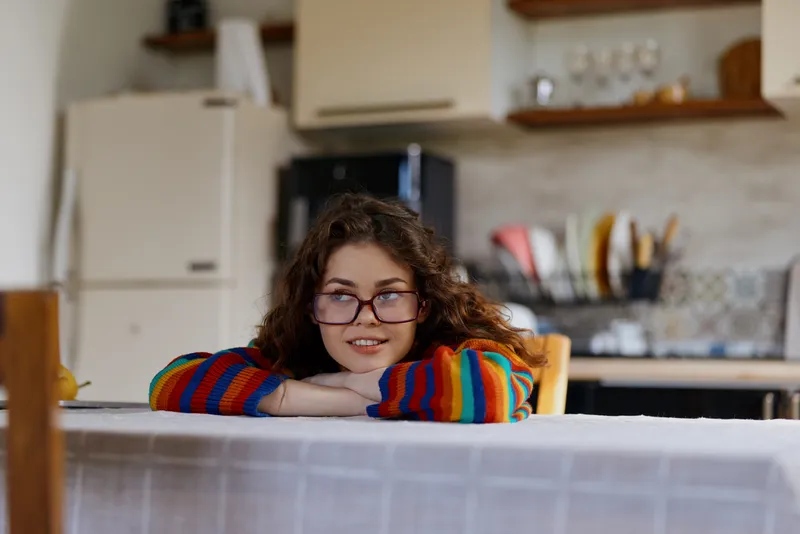 Casual woman indoors in a colorful striped sweater, leaning on a kitchen table with a soft smile, capturing a warm home lifestyle moment in a bright, natural setting