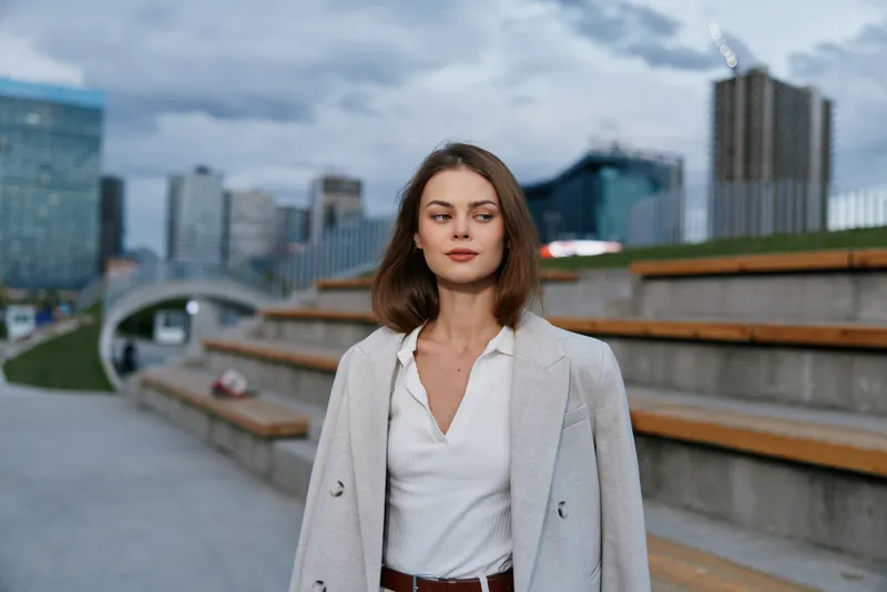 Young woman in white shirt and light coat outdoors with urban cityscape background confident and calm expression KEY business woman, urban, cityscape, confident, calm, young adult, outdoors, coat, cas