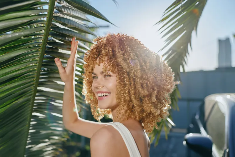 smiling woman with curly hair enjoying sunny day outdoors near tropical palm leaves, wearing white top, bright and vibrant summer atmosphere