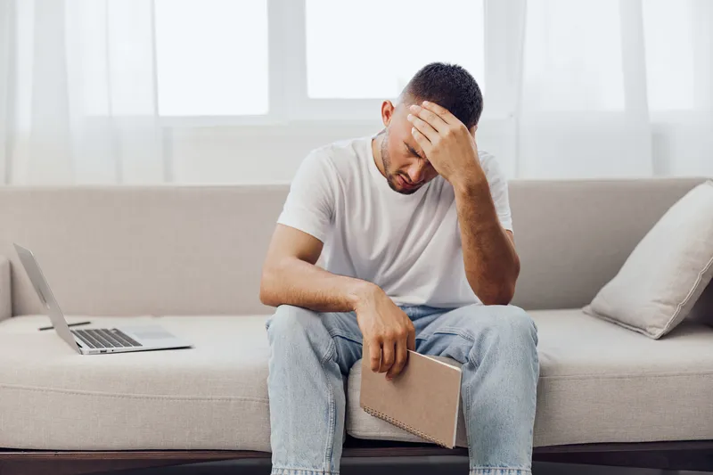 Thoughtful man sitting on a couch, holding a notebook, expressing reflection and contemplation. Modern lifestyle with digital devices in the background.