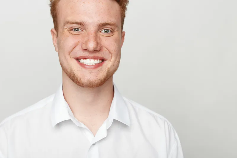 Smiling young man with red hair and beard wearing white shirt looks at camera with bright happy expression. Studio portrait on light background. Concept of confidence, positivity, and youth.