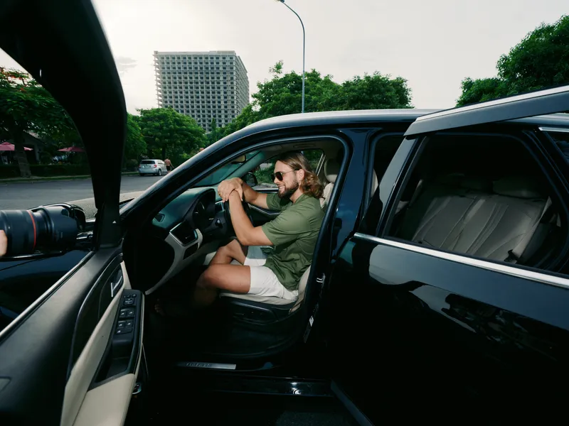 A man sits in the driver seat of an open car with the door ajar, relaxed pose, sunglasses, and a calm expression, set against an urban street and green trees.