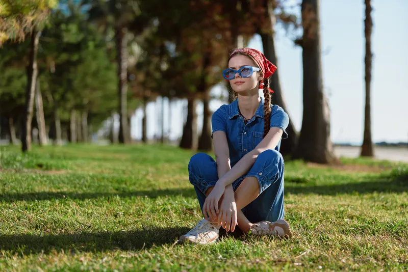 lifestyle woman in modern boho-western denim street style sits relaxed on grass outdoors with film color tones under sunlight and trees in background
