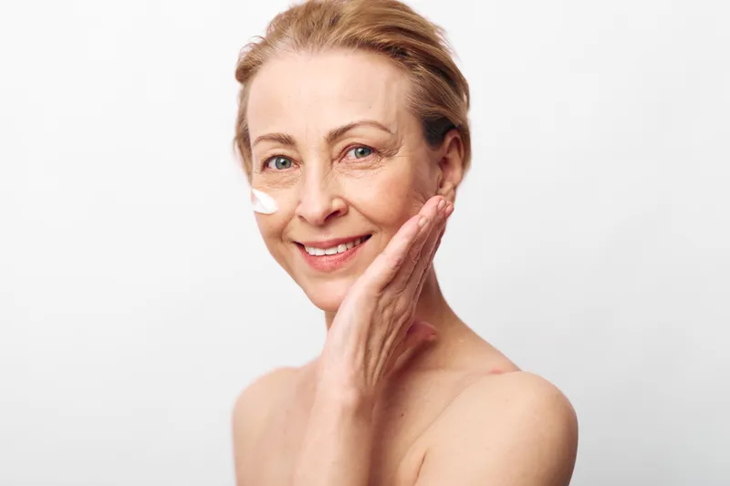 Mature woman with clear skin applies cream on face, smiling gently. Bright studio background highlights natural beauty, skincare, antiaging, and selfcare concepts for healthy skin.