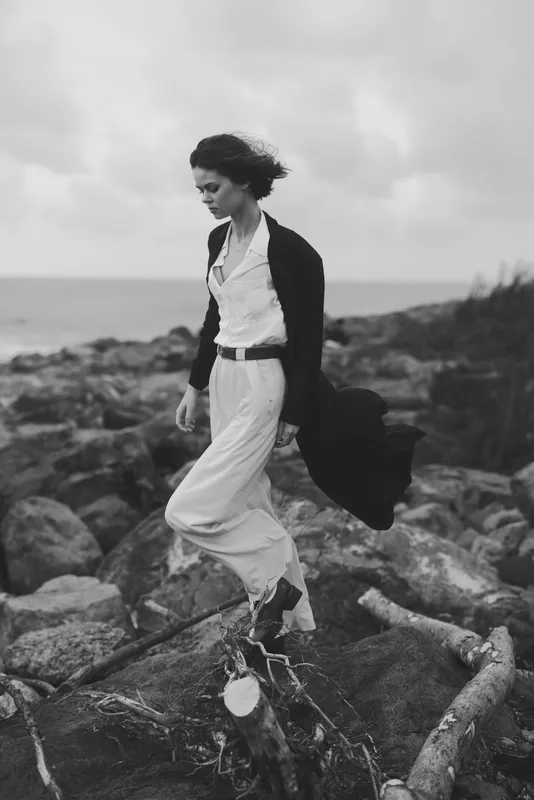 Woman in a white dress and cardigan walks along a rocky shore, holding a coat, as wind and waves frame a moody black and white landscape, conveying solitude, fashion, and resilience against the sea