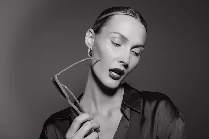 Portrait of a pale skinned white woman with slicked hair holding glasses near her face in black and white studio lighting.