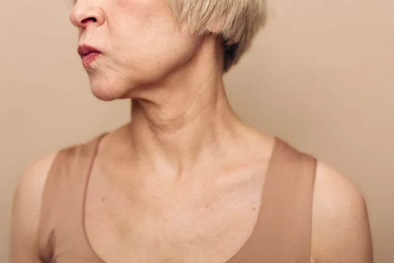 Profile of mature woman with short blond hair and natural skin texture wearing beige top on plain background. Concept of aging, skin care, natural beauty, and lifestyle.