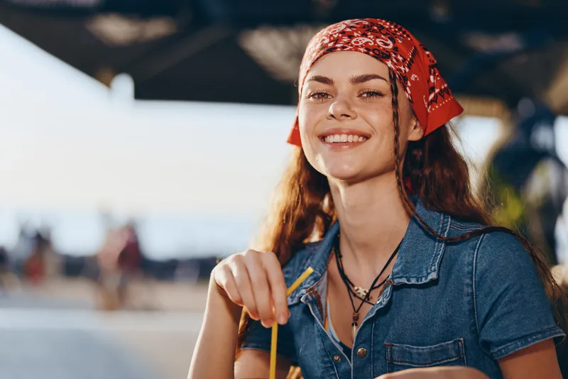 smiling woman enjoying summer drink outdoors with red bandana and denim shirt, natural light, casual style, happy mood, warm atmosphere