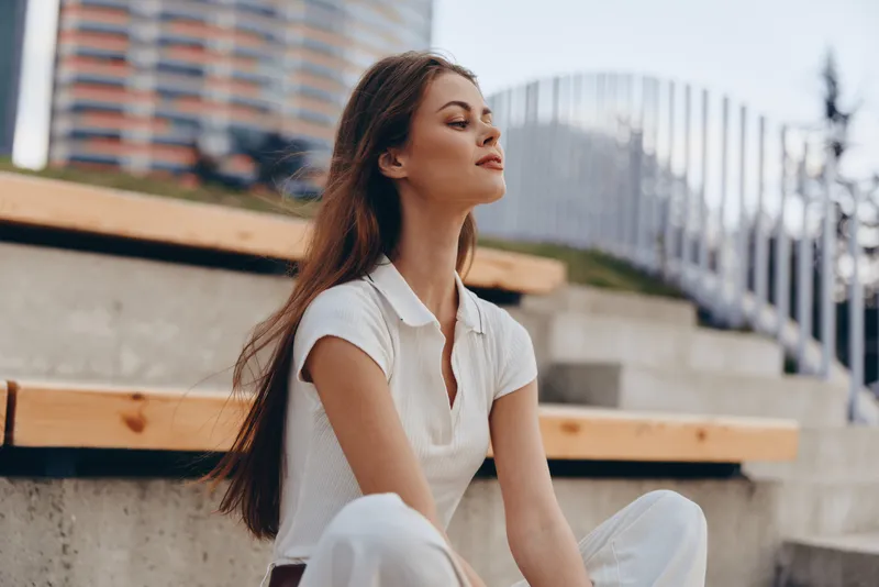 woman enjoying peaceful moment outdoors in casual white outfit with long hair sitting on urban steps during daytime natural light