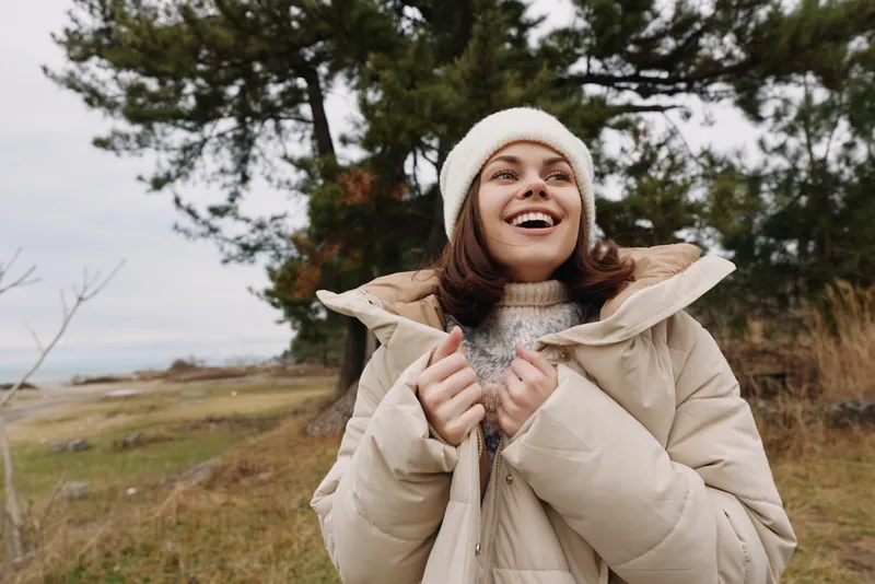Woman in a warm coat and beanie smiles outdoors, standing in a windy landscape with trees and a cloudy sky, conveying fresh air, travel and seasonal mood.