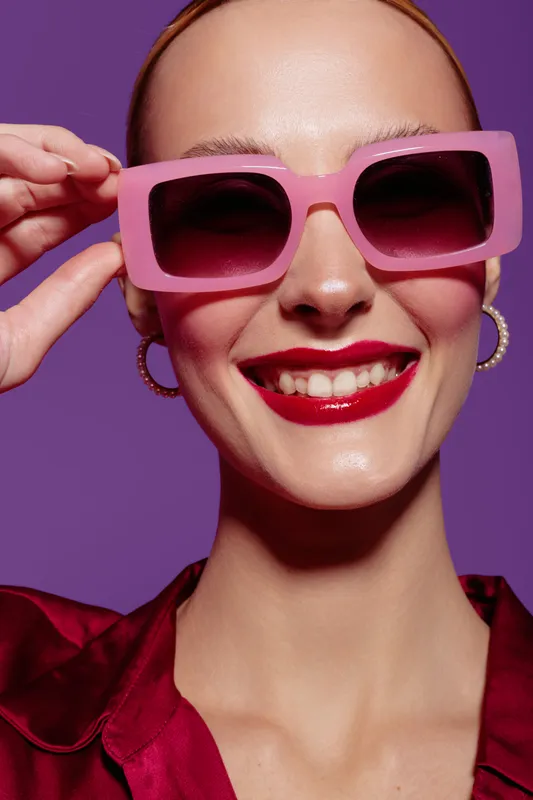 Smiling woman with pink sunglasses and red lipstick poses against purple background. Fashion portrait with bright colors, stylish accessories, and confident expression. Concept of beauty and style.