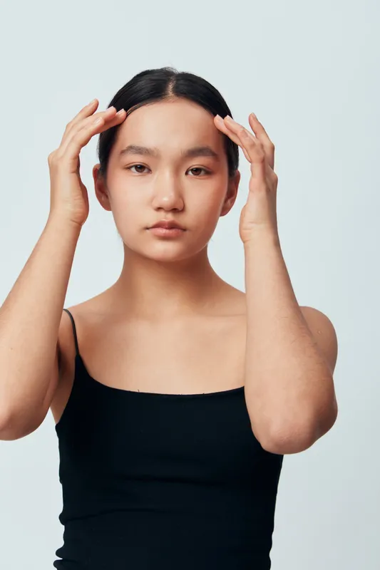 Young woman in black tank top touching face with hands for skincare or beauty routine in minimal studio. Calm expression, natural look and fresh skin with simple background.
