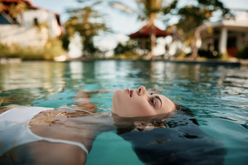 Relaxing woman floats in clear pool water, serene expression, sunny resort backdrop, tropical plants and lounge area, lifestyle scene highlighting leisure, self care, and peaceful moment