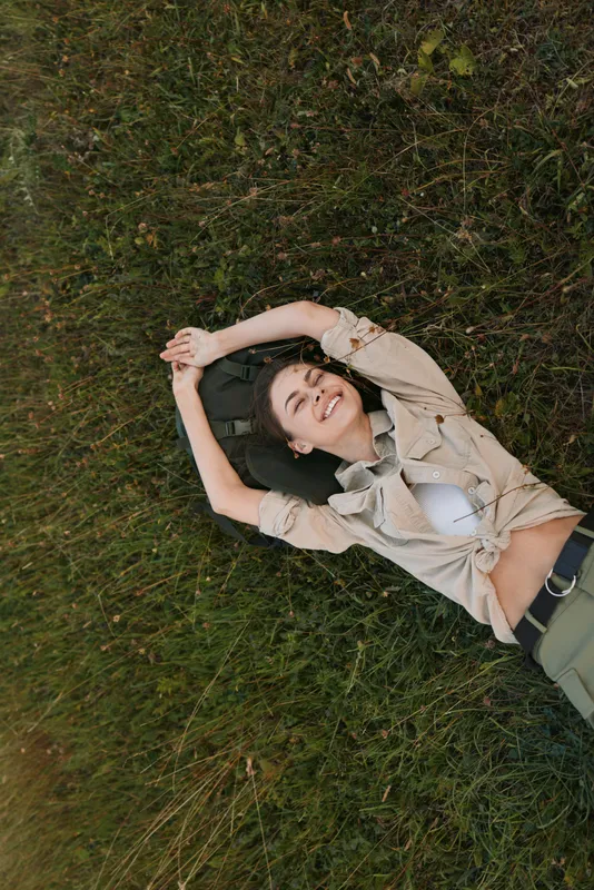 Young woman lies on grass in a relaxed outdoor scene, wearing a beige jacket over a white top with green pants, smiling softly as a gentle breeze blows.
