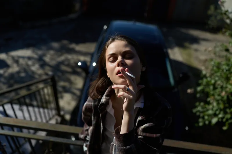 Young woman on a sunny balcony smokes a cigarette with closed eyes, relaxed posture, urban background and warm light creating a casual lifestyle moment